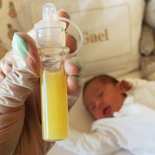 Load image into Gallery viewer, Colostrum collector with yellow liquid (colostrum breastmilk) held by a gloved hand, baby lying in crib in the background
